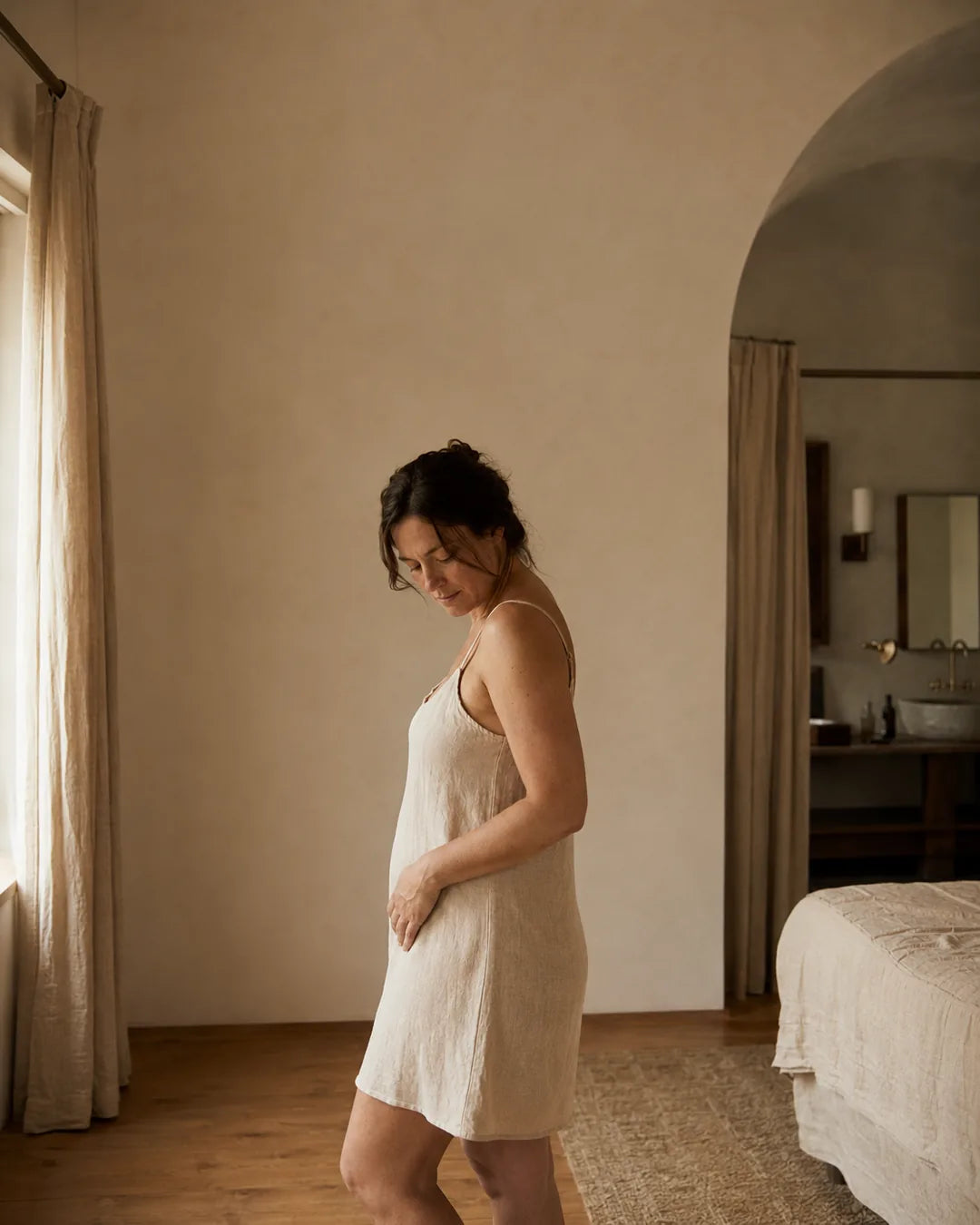 A woman in a linen slip in soft afternoon light, looking down and considering her body in a boutique bedroom.